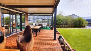 A covered patio with outdoor furniture, a herringbone wooden floor, and a view of a green lawn and water body.