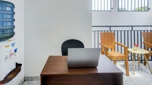 Una habitación con una mesa de madera, una computadora portátil, sillas de mimbre, un dispensador de agua y vistas al exterior a través de una ventana con persianas verticales.