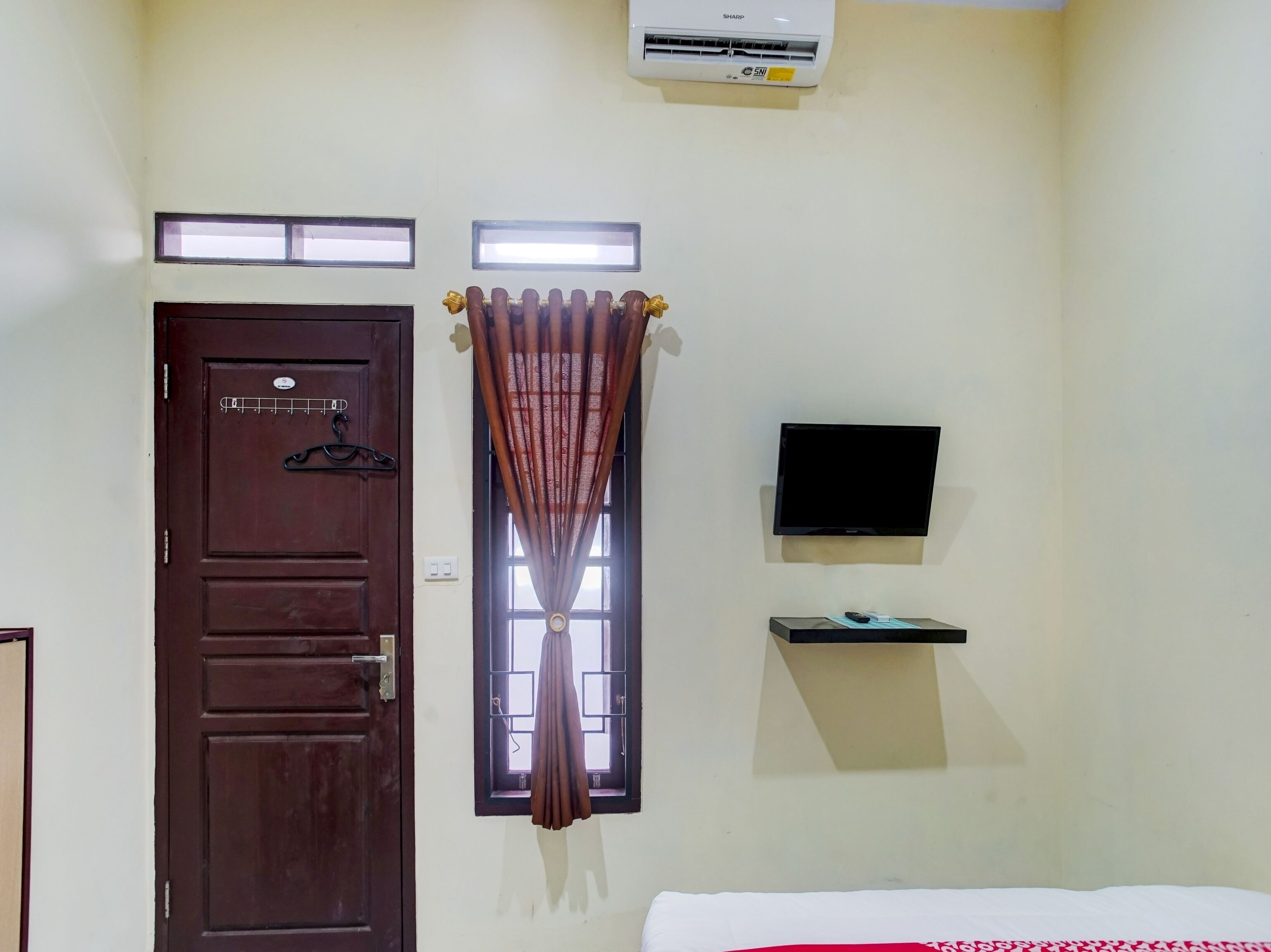 Quarto com porta marrom, janela com cortinas, televisão montada e uma pequena prateleira.