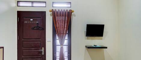 Una habitación con una puerta marrón, una ventana con cortinas, un televisor montado y un estante pequeño.