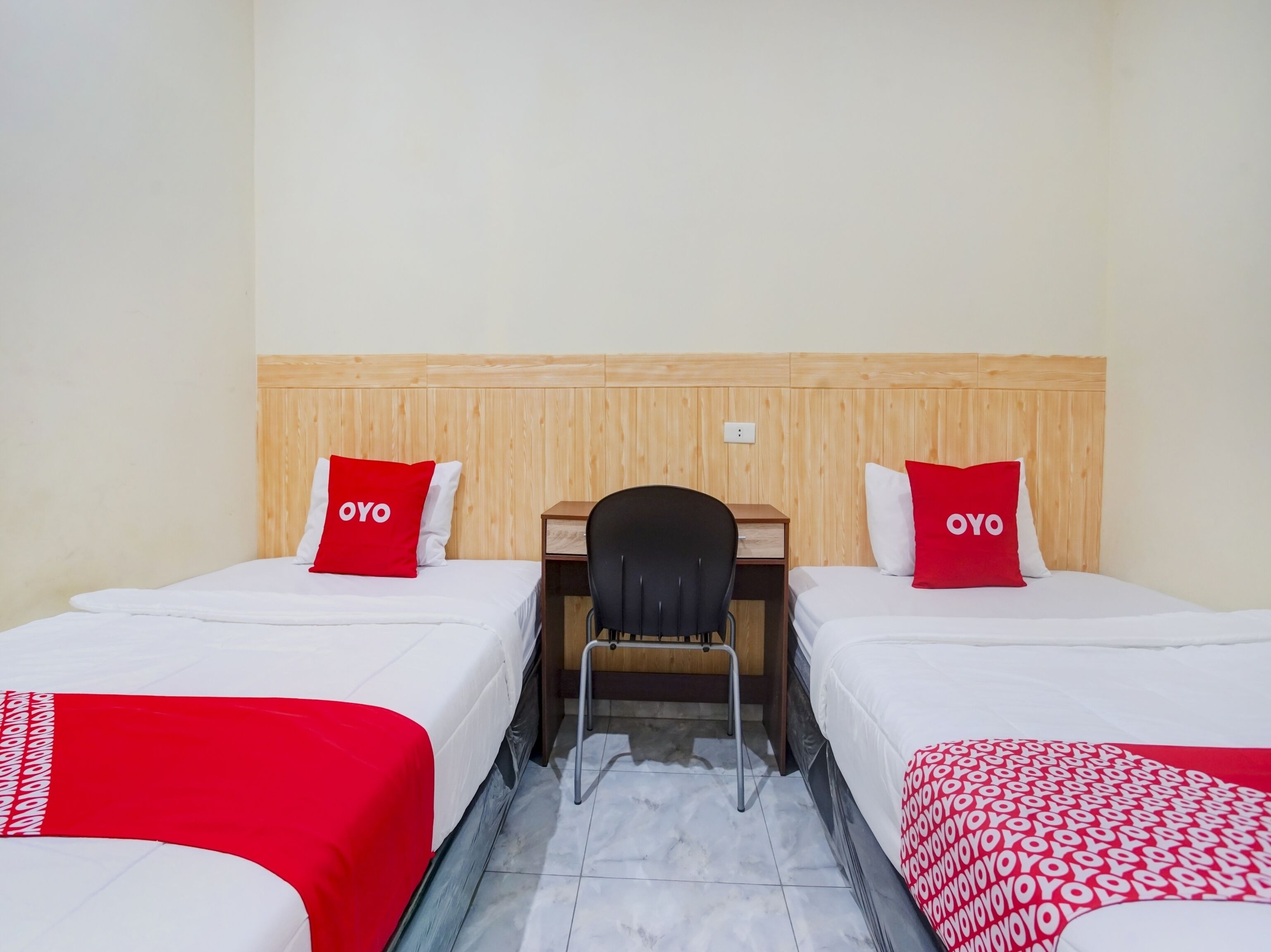 Una habitación de hotel con dos camas individuales, cada una con un almohadón rojo con la etiqueta "OYO", un cabecero de madera, un escritorio con una silla y piso de baldosas.