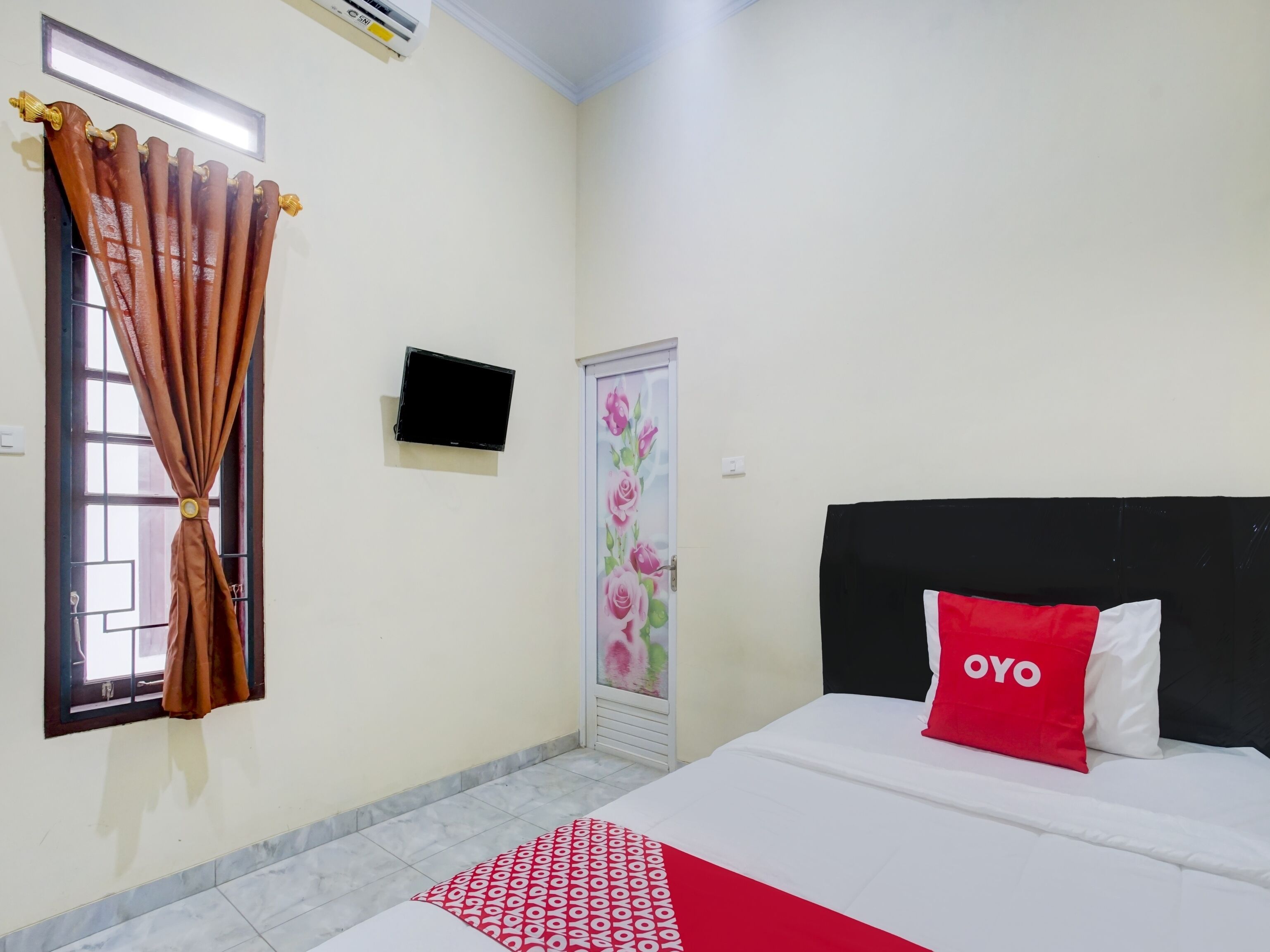 Quarto de hotel com cama, televisão, janela com cortinas e porta com detalhe floral.