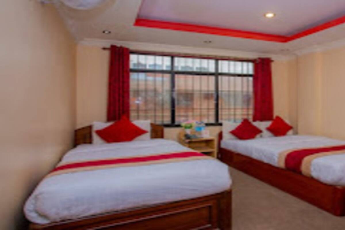 A hotel room with two beds, red pillows, a small table, and a window with curtains.