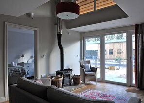 Ein modernes Wohnzimmer mit Kamin, einer Couch, einem Sessel und einem großen Fenster mit Blick ins Freie.