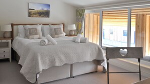 A bedroom with a large bed, two bedside tables, a window with wooden blinds, and a view of a building outside. - Modern Barn Conversion, 3 bedrooms (Cheltenham)