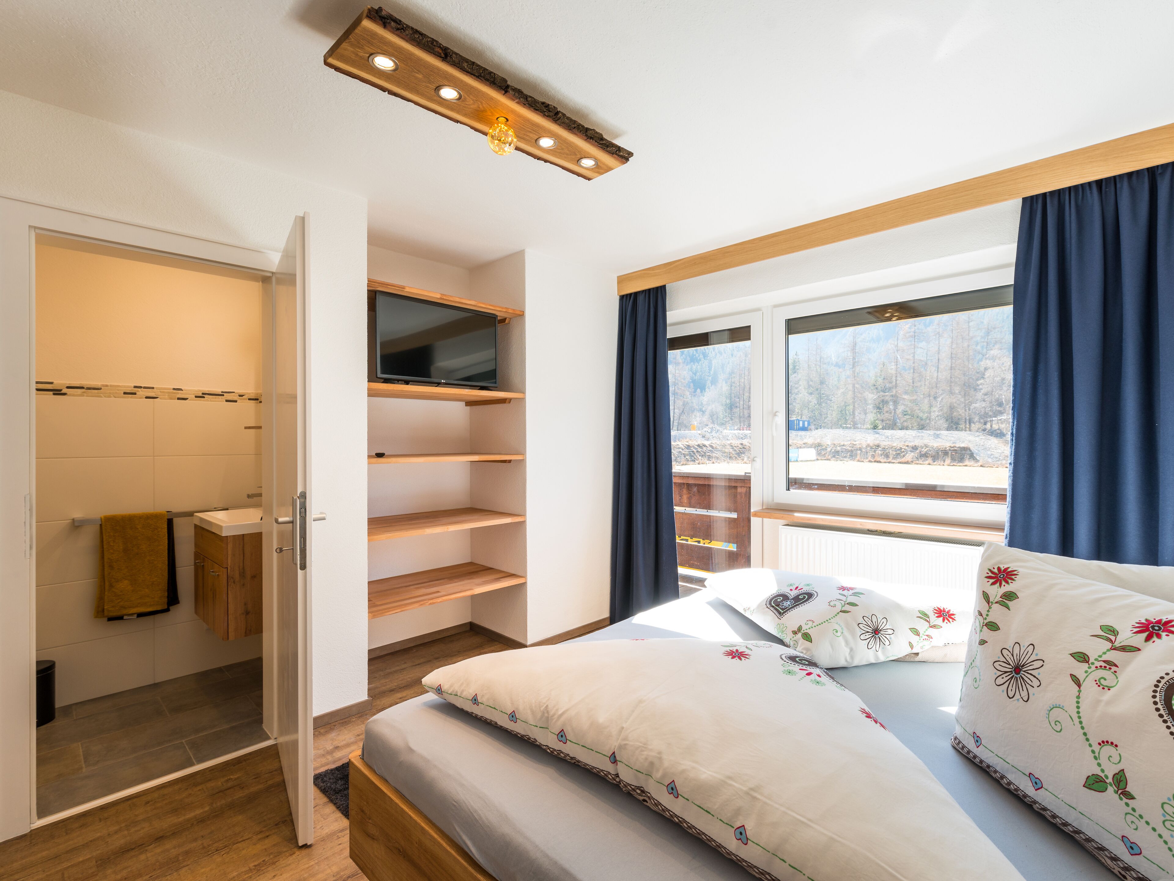 Une chambre à coucher avec un lit, un téléviseur fixé sur une étagère en bois, une fenêtre avec des rideaux et une vue sur un paysage enneigé.