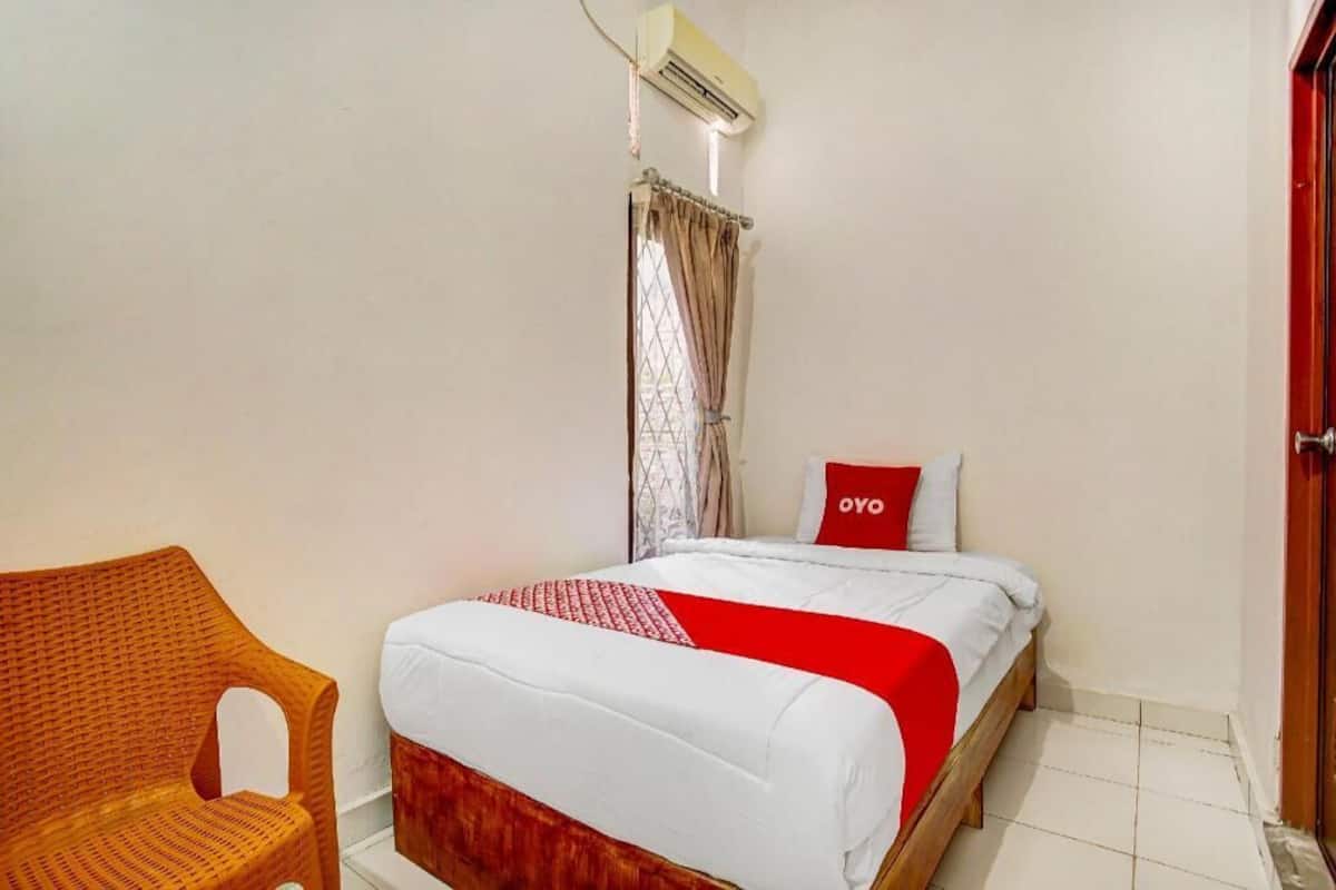 A single bed with a red and white bedspread, a wooden headboard, a wicker chair, a window with curtains, and an air conditioning unit.