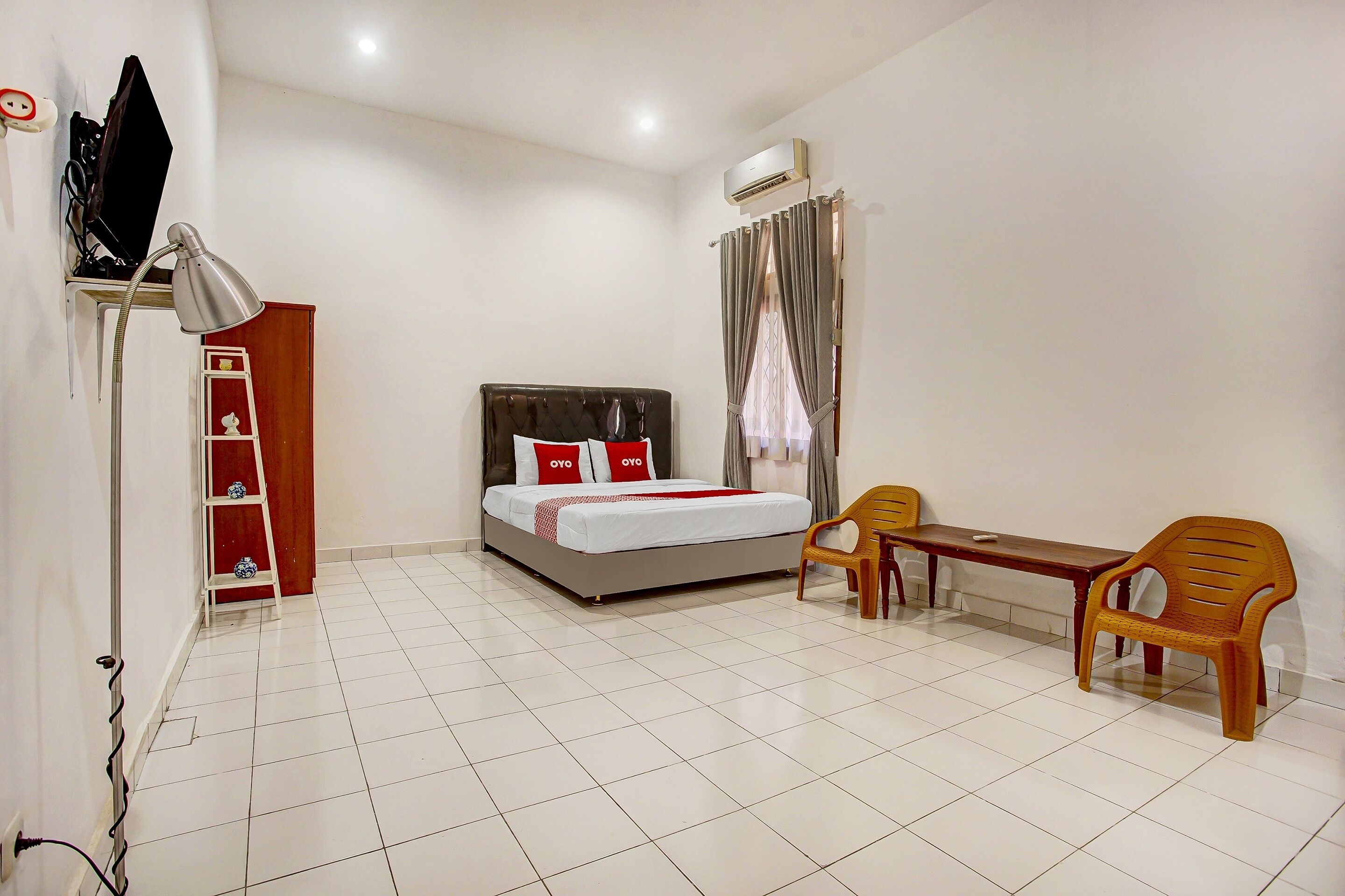 A hotel room with a bed, a desk, a chair, a shelf with decorative items, and an air conditioning unit.