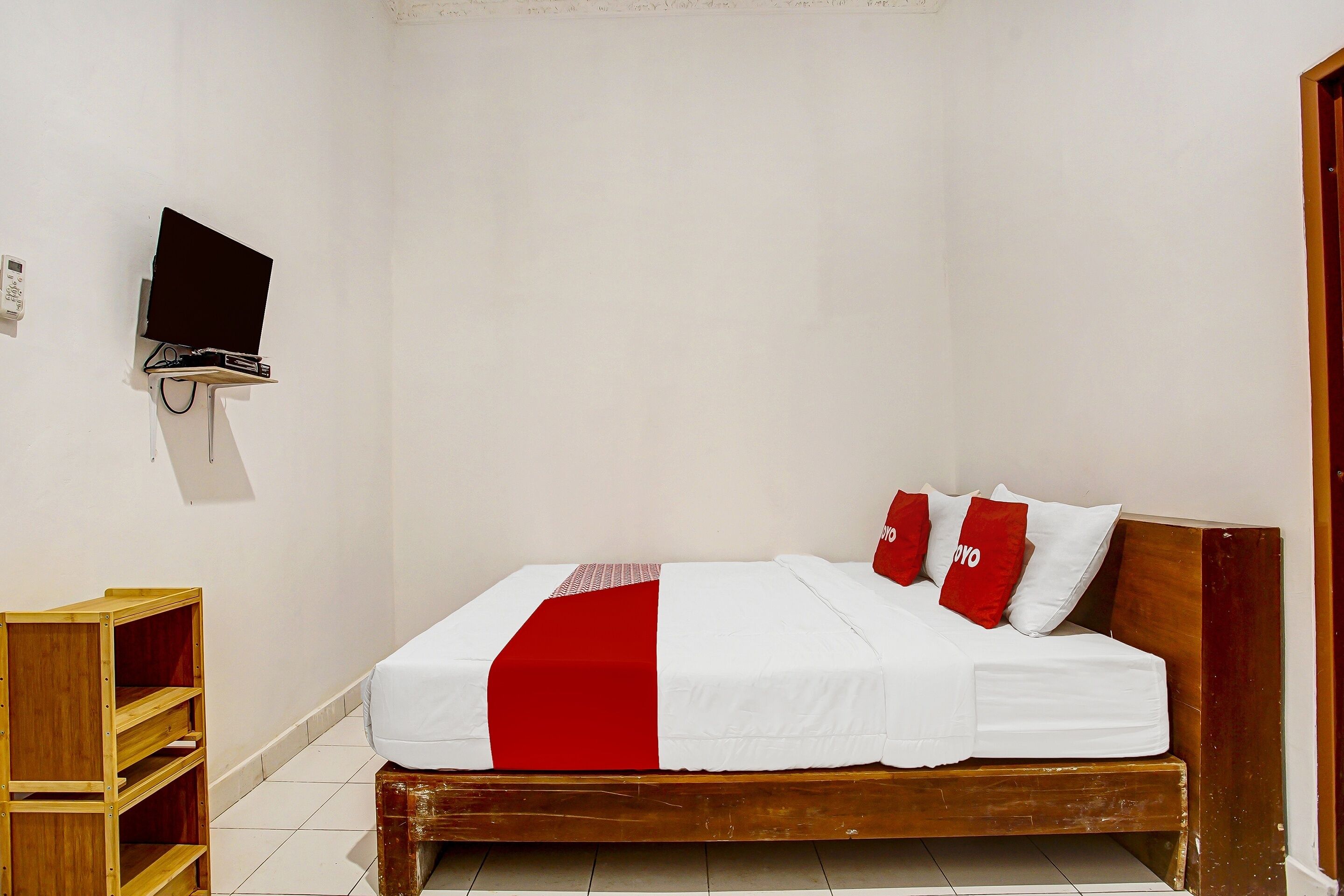 Un lit simple avec un cadre en bois, une literie blanche et des touches de couleur rouge. Un téléviseur est fixé au mur au-dessus du lit.