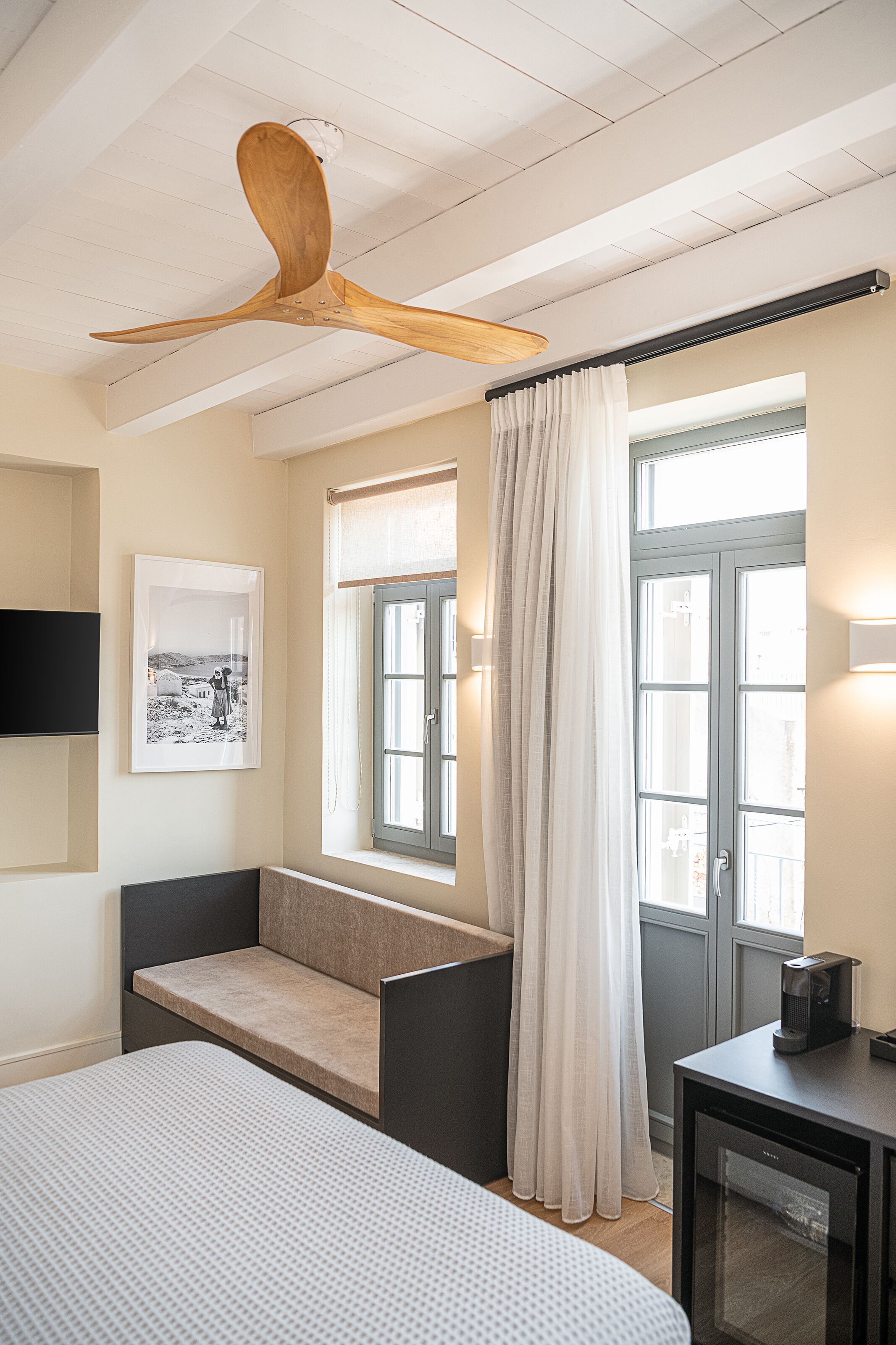 Une chambre à coucher avec un lit, un ventilateur de plafond en bois, une fenêtre avec des rideaux, une table de chevet et un téléviseur fixé au mur.
