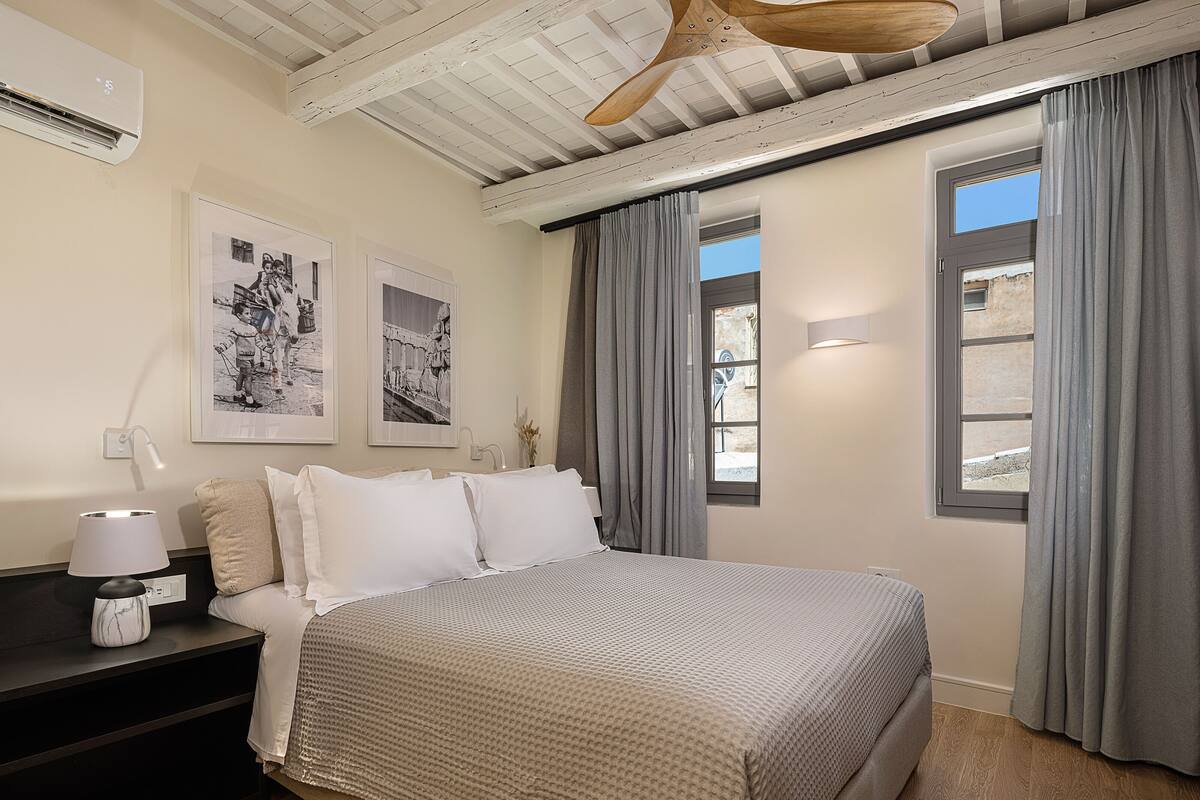 A bedroom with a bed, a nightstand, a lamp, and two windows with curtains.