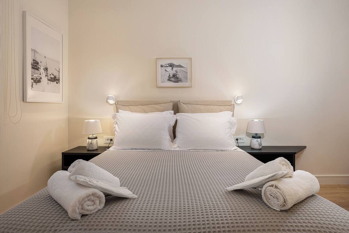 A neatly made bed with white linens and two rolled towels, a bedside table with a lamp, and a framed picture on the wall.