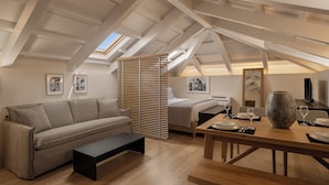 Un acogedor dormitorio tipo loft con techo inclinado, una cama, un sofá, una mesa de comedor con un florero y cuadros enmarcados en las paredes.
