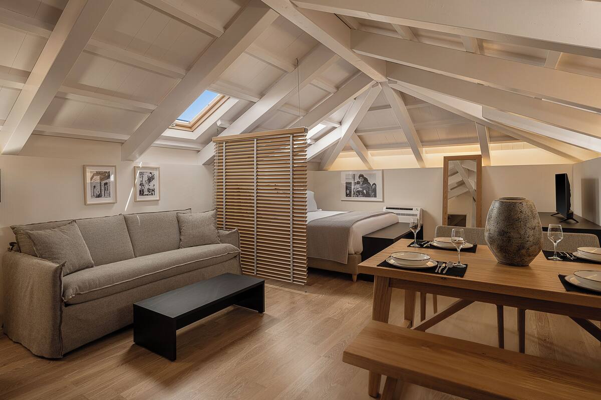A cozy loft bedroom with a sloped ceiling, a bed, a sofa, a dining table with a vase, and framed pictures on the walls.