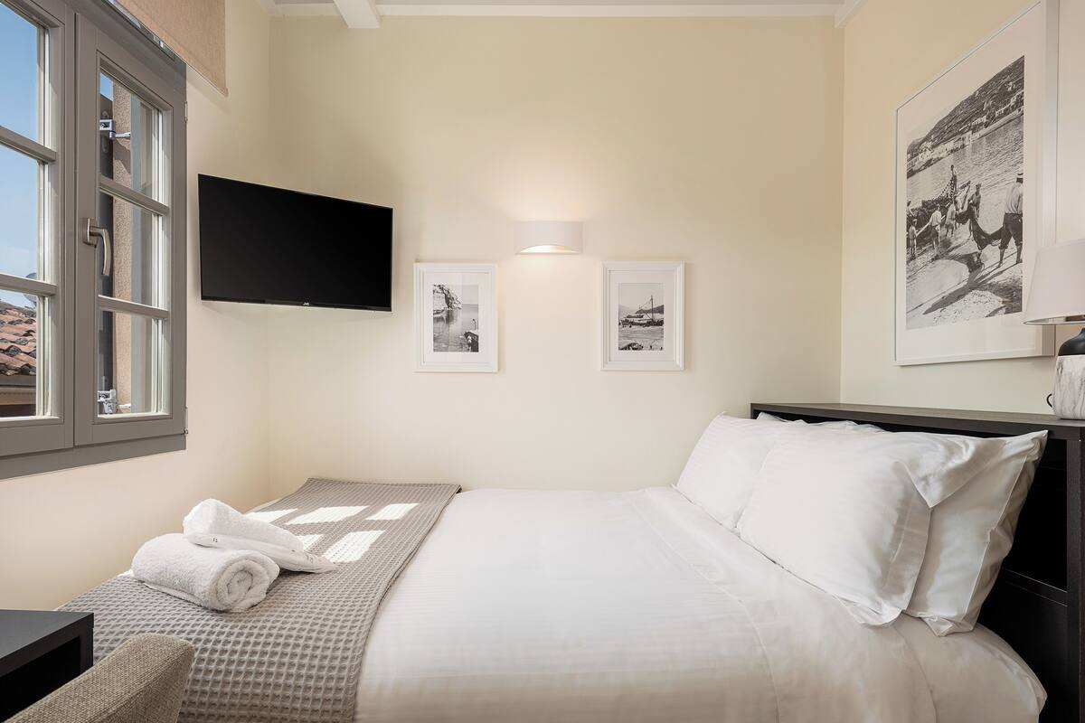 A neatly made bed with white linens, a TV mounted on the wall, and two framed pictures on the wall.