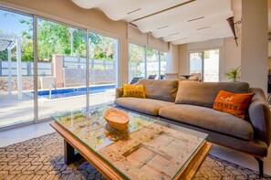 A modern living room with a glass coffee table, a grey sofa with 'HERMES' pillows, and a view of a pool and outdoor area.
