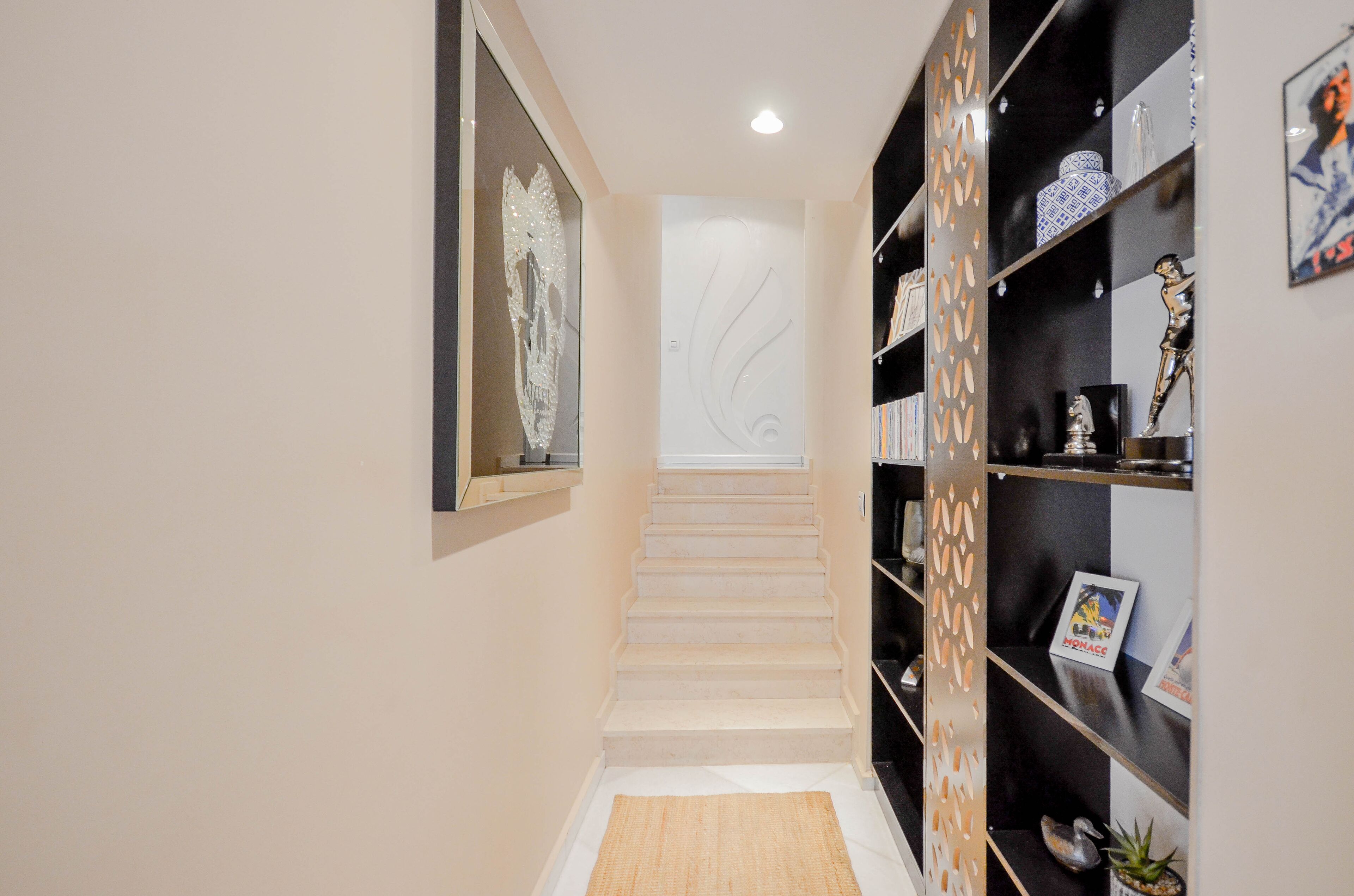 Un pasillo con una escalera, un muro decorativo y una estantería con varios objetos.