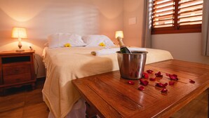Une chambre d’hôtel avec un lit, des lampes de chevet, une table en bois sur laquelle se trouvent une bouteille et des pétales de rose, et une fenêtre avec des stores.