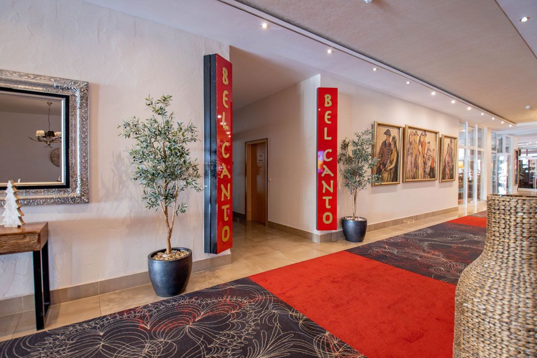 A hotel lobby with red pillars labeled "BELCANTO," a large mirror, potted plants, and framed artwork on the walls.