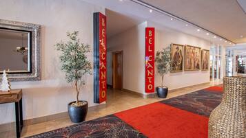 A hotel lobby with red pillars labeled "BELCANTO," a large mirror, potted plants, and framed artwork on the walls.