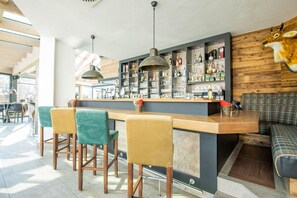 Una zona de bar con mostrador de madera, taburetes y estantes llenos de botellas y vasos.