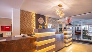 A modern reception desk with a brick-patterned wall, a clock, and a chandelier. - Hotel Tanne (Ilmenau)