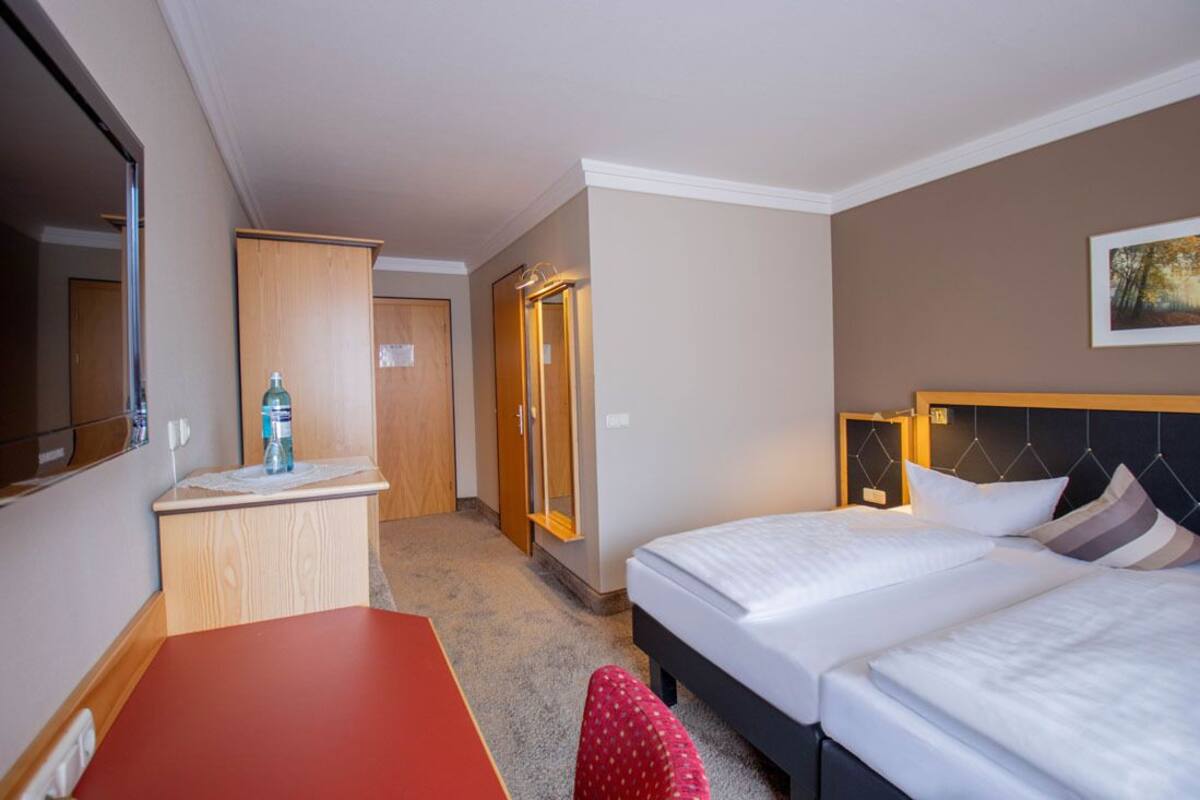 A hotel room with two beds, a desk, a mirror, a wardrobe, and a painting on the wall.