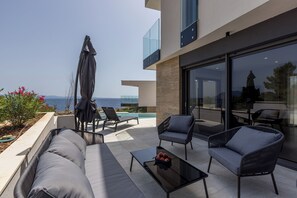 A modern outdoor seating area with a pool, lounge chairs, and a glass balustrade overlooking the sea.
