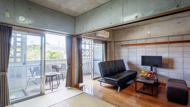 A modern living room with a black leather sofa, a small table with a lamp, and a view of the city through large sliding glass doors. - Nago Parkside Condominium TK Stay (Nago)