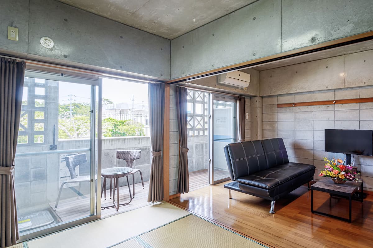 A modern living room with a black leather sofa, a small table with a lamp, and a view of the city through large sliding glass doors.