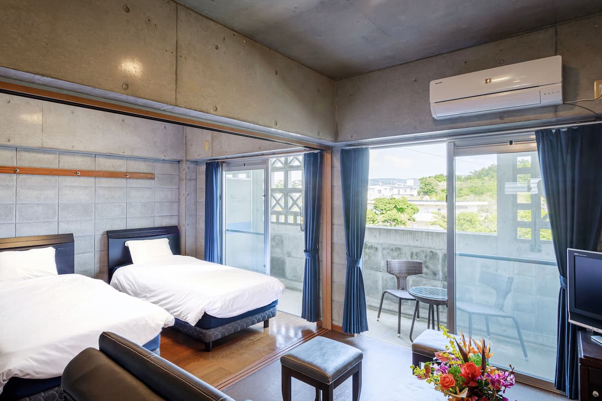 A hotel room with two beds, a TV, a balcony with chairs, and an air conditioning unit.
