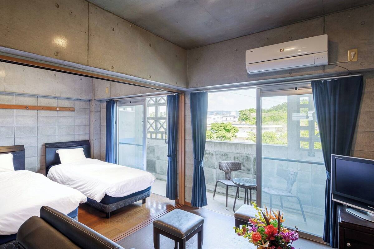 A hotel room with two beds, a balcony with a TV, and an air conditioning unit.