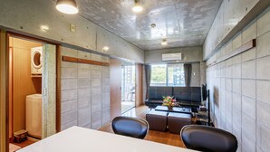 A modern hotel room with a large window, a sofa, a dining table, and a washing machine. - Nago Parkside Condominium TK Stay (Nago)