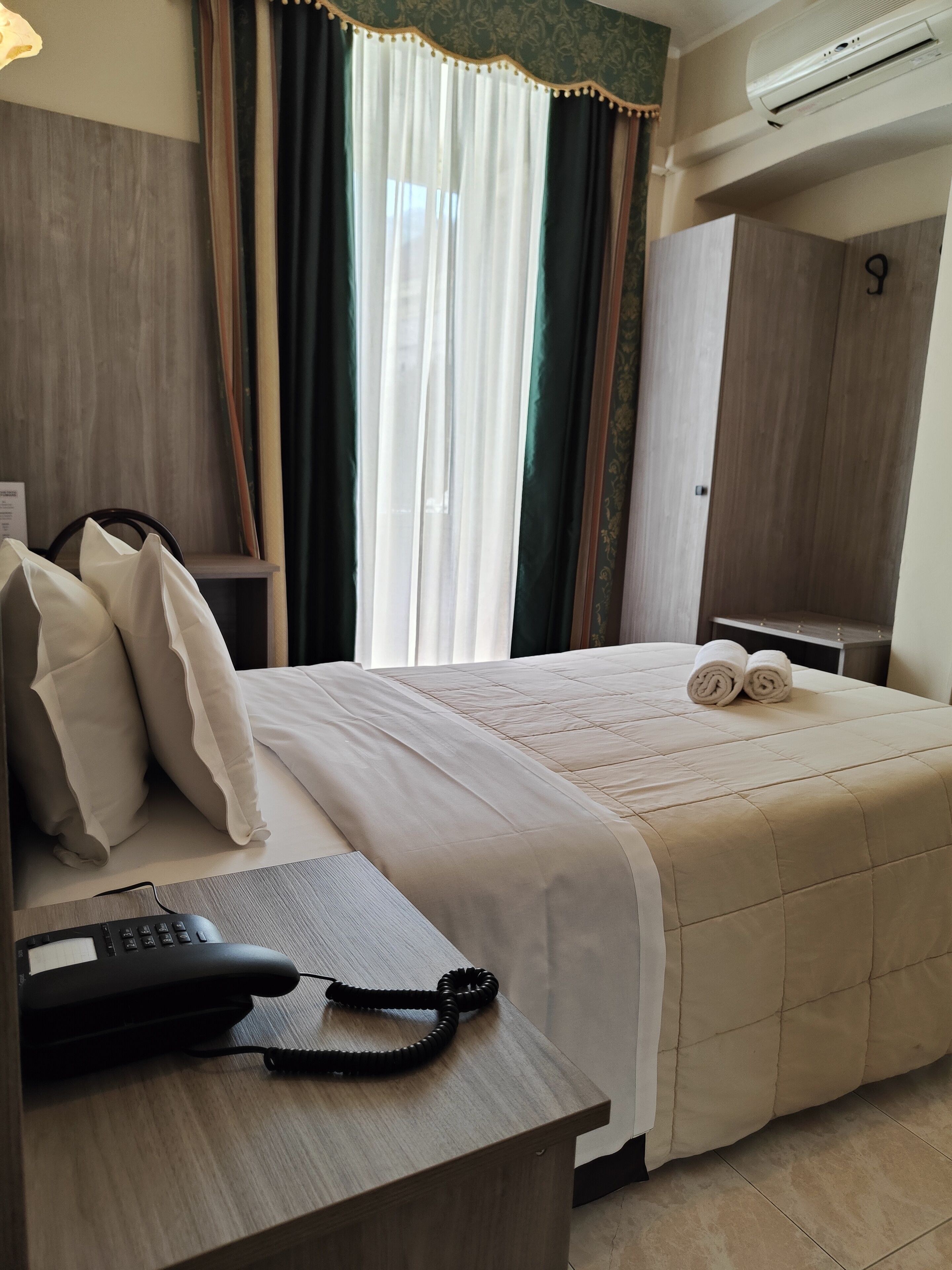 A hotel room with a bed, a desk with a telephone, a wardrobe, and an air conditioning unit.