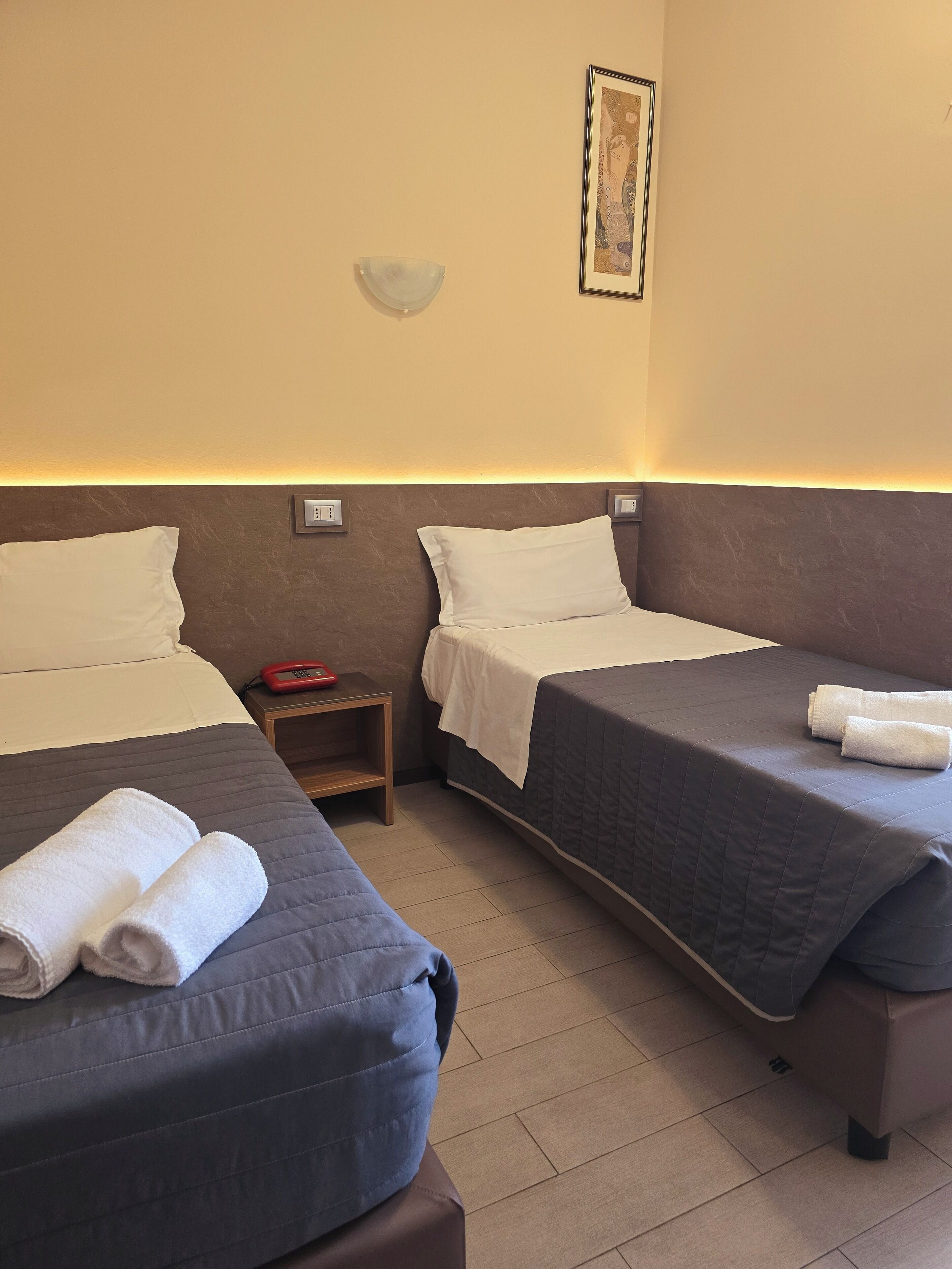 Camera d'albergo con due letti, ognuno con cuscino e coperta, un comodino con una sveglia rossa e un lampadario a parete.