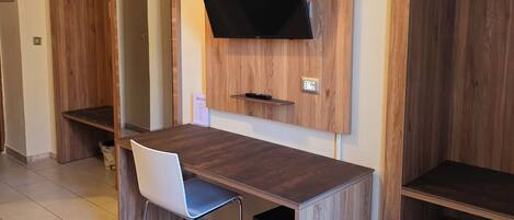 A hotel room with a wooden desk, a chair, a flat-screen TV mounted on the wall, and a small shelf below the TV.