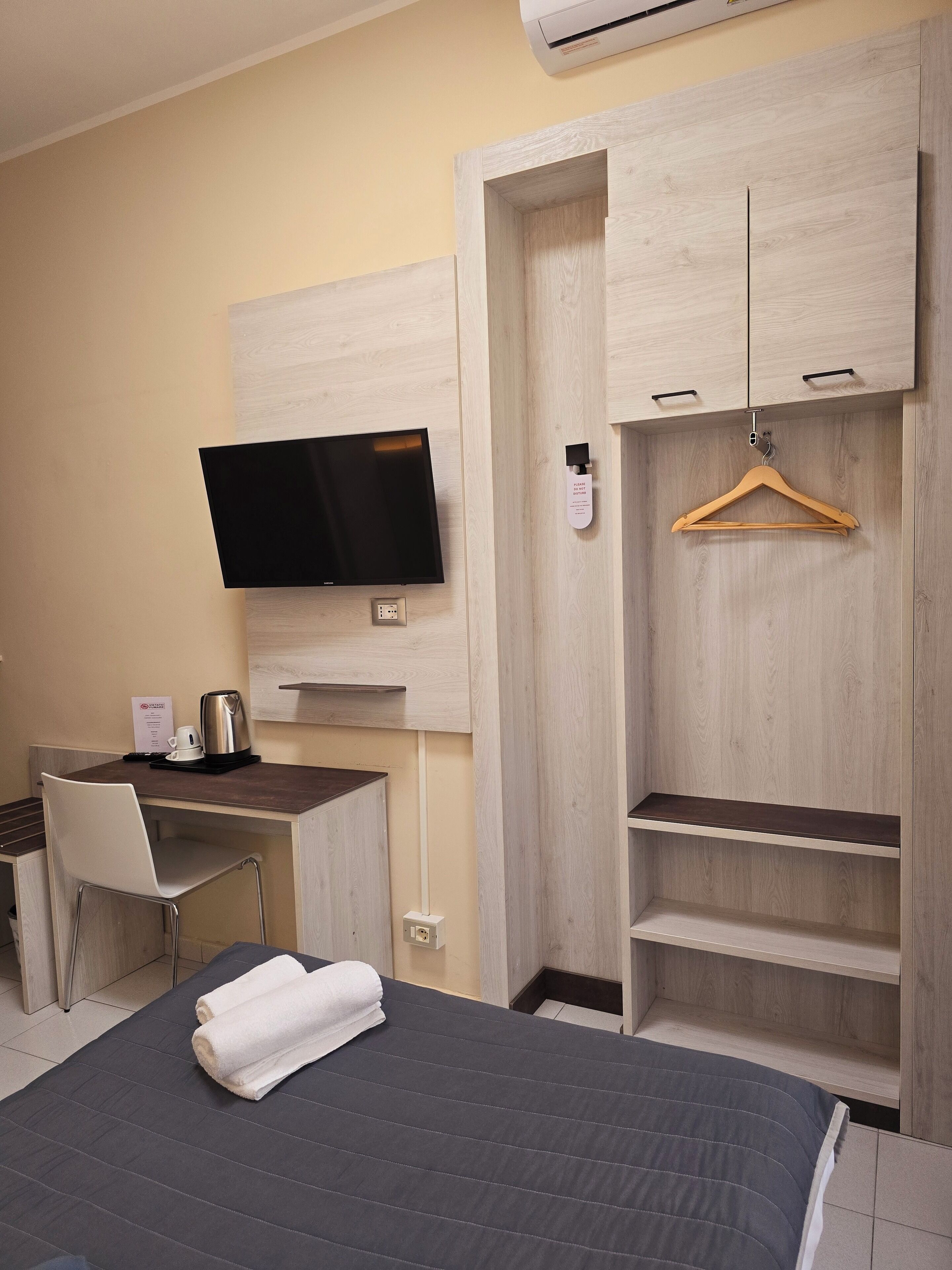 Quarto de hotel com cama, escrivaninha com cadeira, televisão e guarda-roupa com cabideiro.