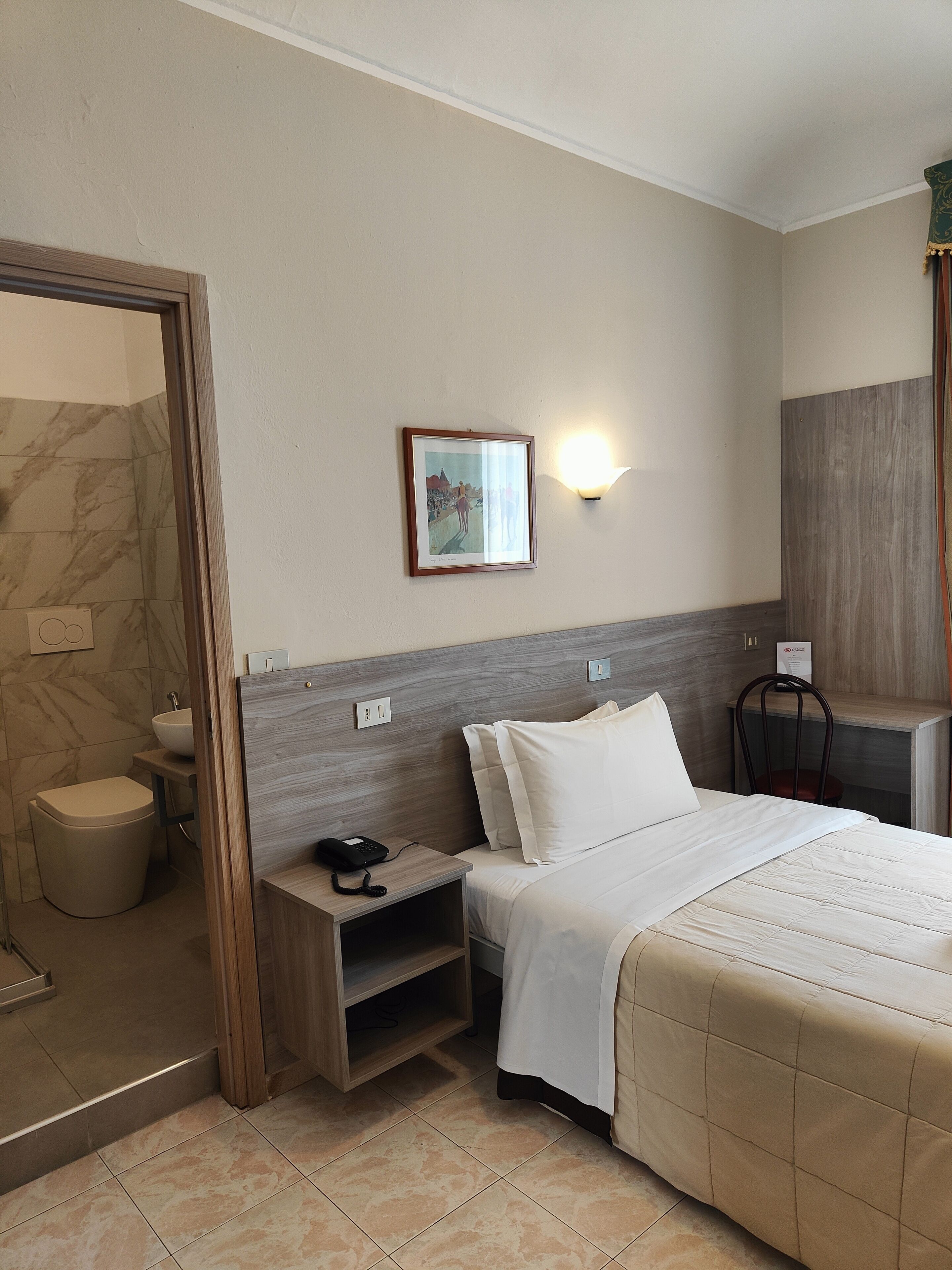 A hotel room with a bed, bedside table, nightstand, and a wall-mounted light fixture.