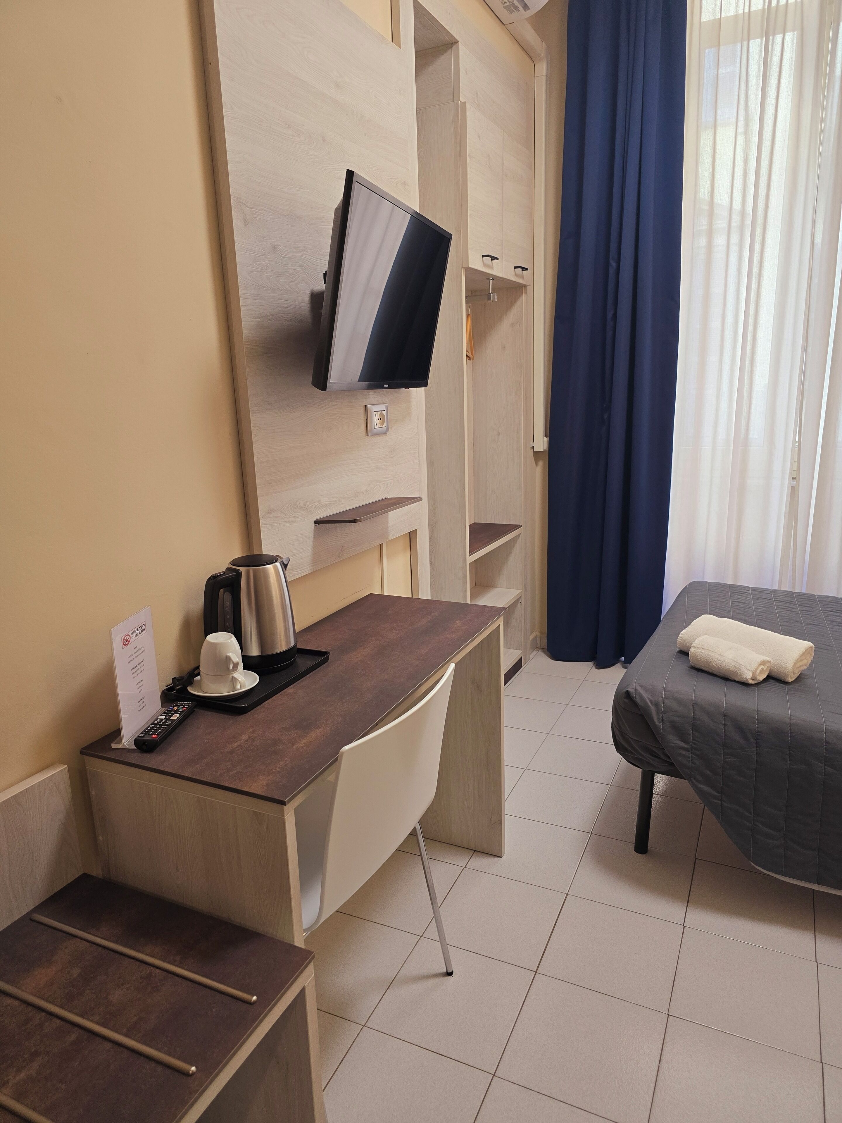 Ein Hotelzimmer mit Schreibtisch, einem Bett, einem an der Wand befestigten Fernseher, einer Kaffeemaschine und einer Fernbedienung auf dem Schreibtisch.
