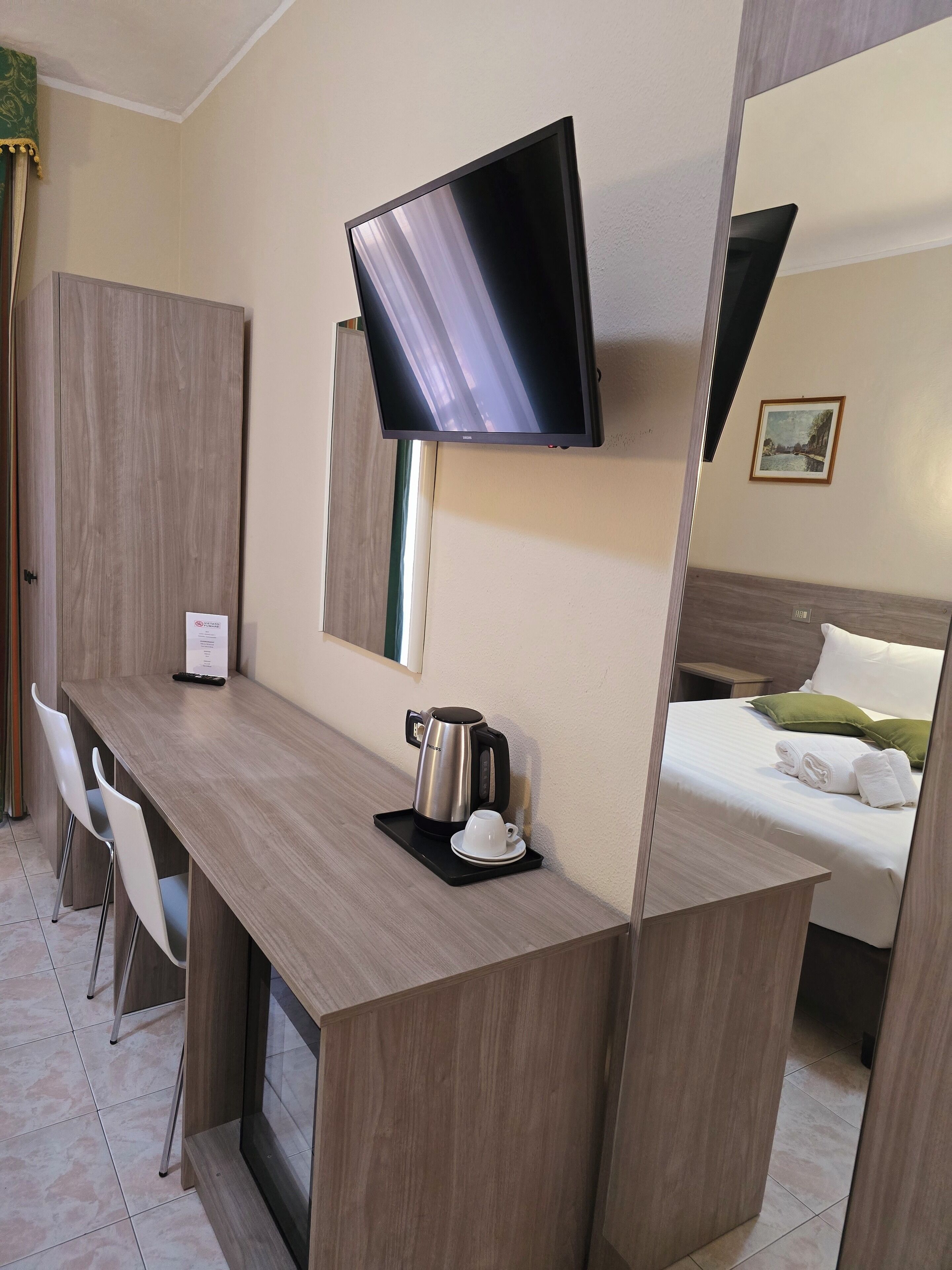 A hotel room with a wooden desk, a flat-screen TV mounted on the wall, a bed with white linens and green pillows, and a coffee maker on the desk.