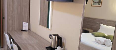 A hotel room with a wooden desk, a flat-screen TV mounted on the wall, a bed with white linens and green pillows, and a coffee maker on the desk.