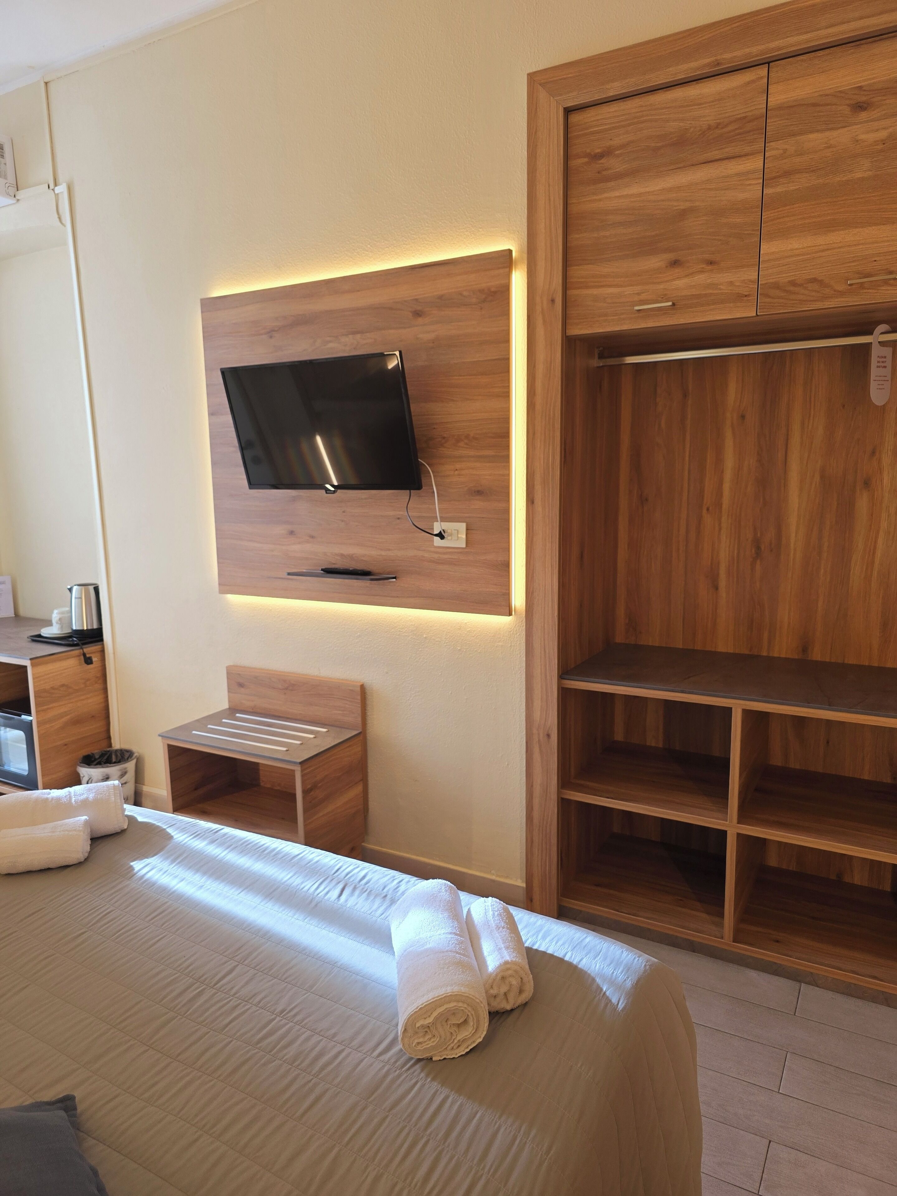 A hotel room with a bed, a TV mounted on the wall, a wooden wardrobe, and a small table with a chair.
