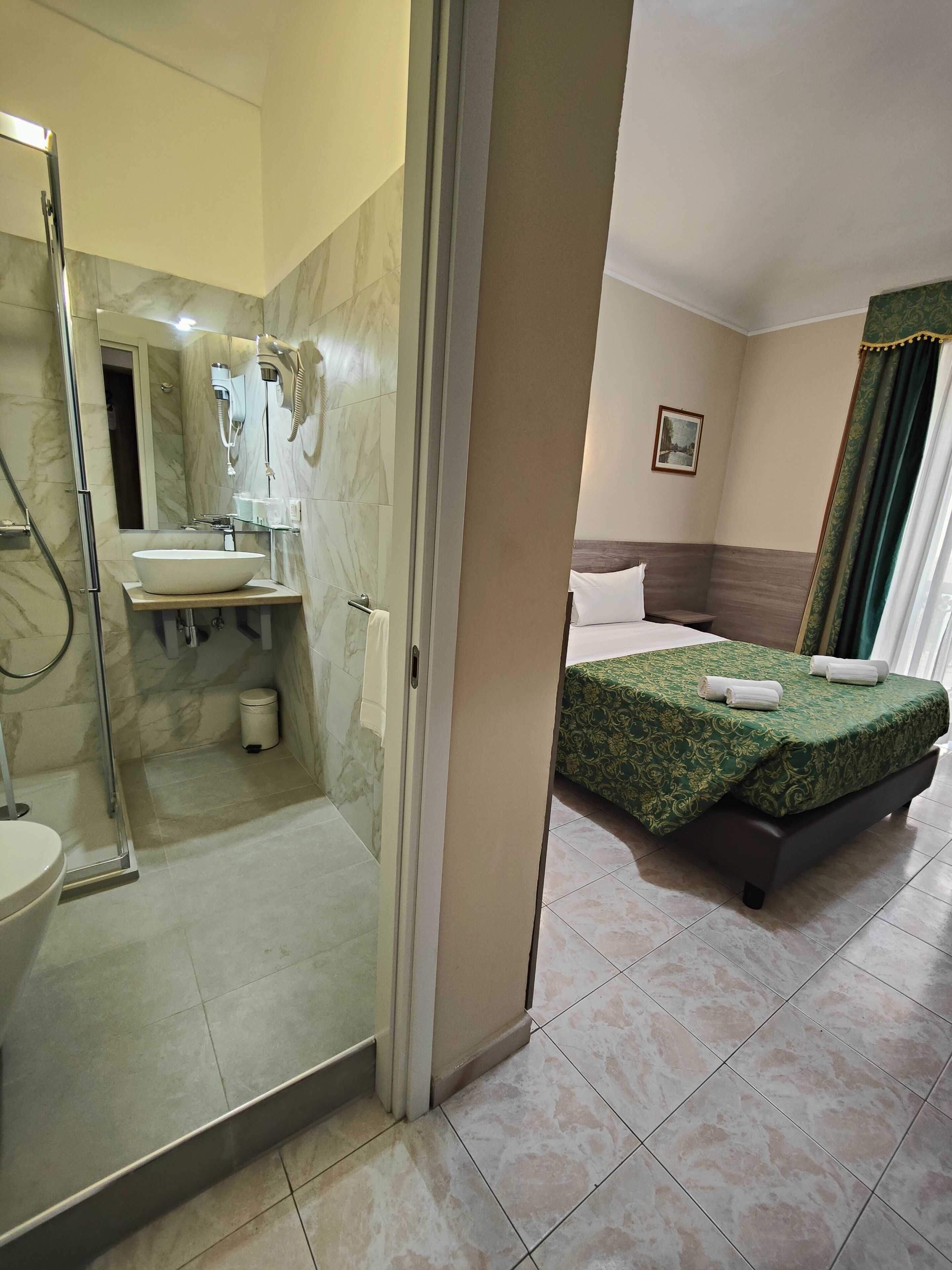 A hotel room with a bathroom featuring a glass-enclosed shower, a sink, and a toilet. The room has a bed with green bedding and a painting on the wall.