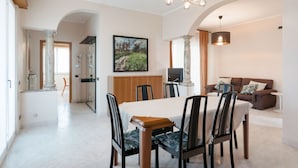 Un'ampia sala da pranzo con un grande tavolo e sedie, un lampadario moderno, e un accogliente angolo salotto con un divano e un quadro appeso al muro.
