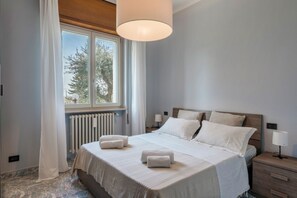 Ein Schlafzimmer mit einem großen Bett, weißer Bettwäsche, einem Nachttisch mit Lampe und einem Fenster mit Blick auf Bäume.