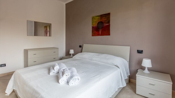 A neatly made bed with white linens and rolled towels, a bedside table with a lamp, a dresser, and a wall-mounted painting. - GARDASUN 2 - Lovely flat Lake close Pet friendly (Padenghe Sul Garda)