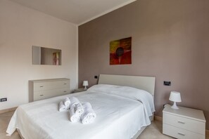 A neatly made bed with white linens and rolled towels, a bedside table with a lamp, a dresser, and a wall-mounted painting. - GARDASUN 2 - Lovely flat Lake close Pet friendly (Padenghe Sul Garda)