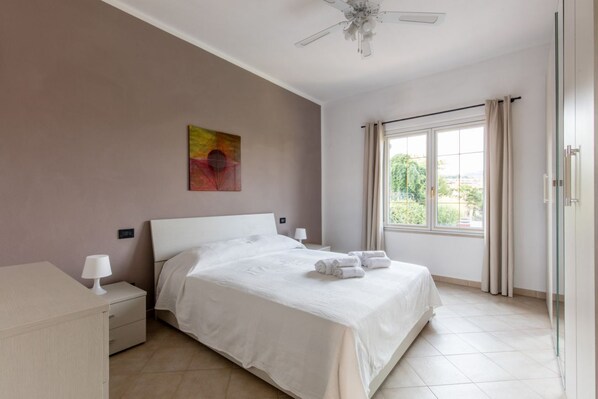 A modern bedroom with a large bed, a bedside table with a lamp, a ceiling fan, and a window with curtains. - GARDASUN 2 - Lovely flat Lake close Pet friendly (Padenghe Sul Garda)