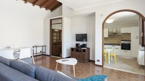 Een moderne woonkamer met een bank, een klein tafeltje en een televisie. Er is een eethoek met een witte tafel en gele stoelen. De keuken is zichtbaar door een boogvormige doorgang.