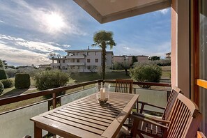 Un balcón con una mesa y sillas de madera, un florero de vidrio con velas y vistas a un edificio de varios pisos y vegetación.